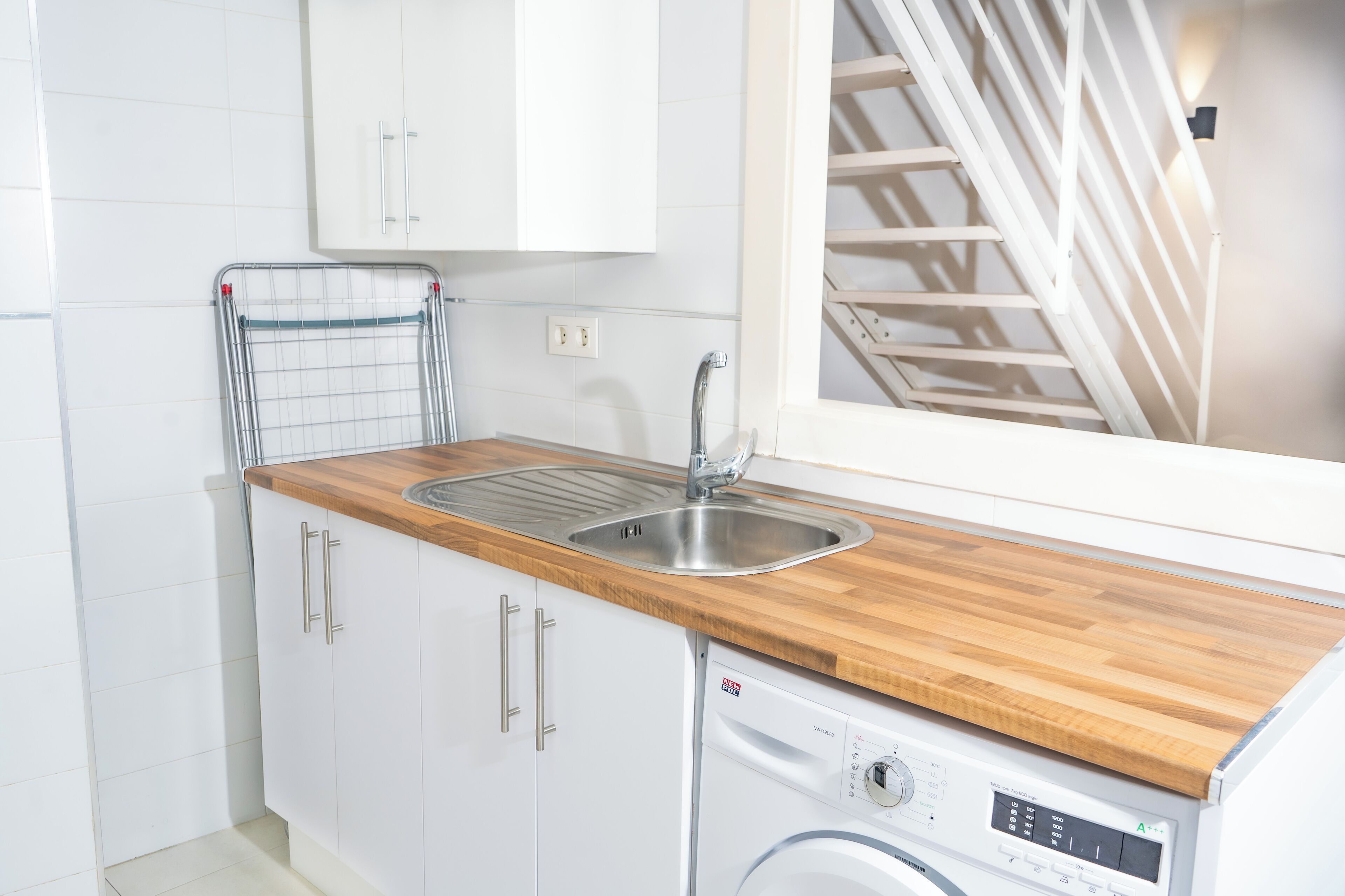 Una cocina con armarios blancos, una encimera de madera, un lavamanos de acero inoxidable y una lavadora integrada.
