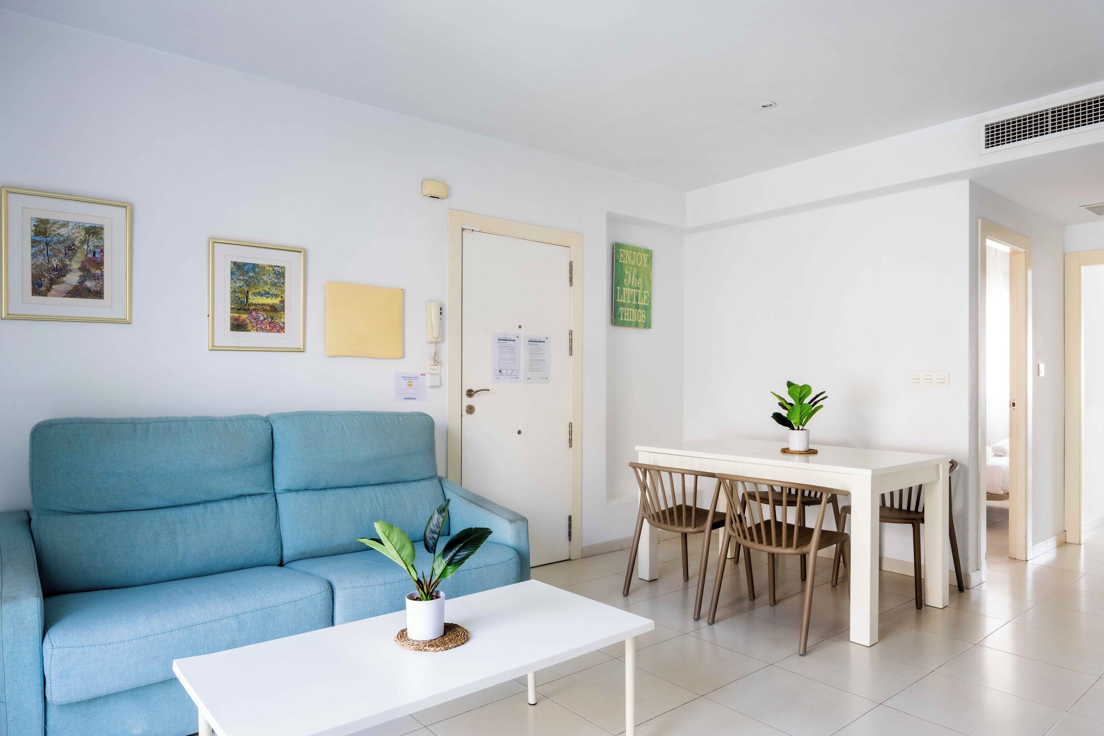 Una sala de estar moderna con un sofá azul, una mesa de centro blanca y una zona de comedor con una mesa blanca y sillas de madera.