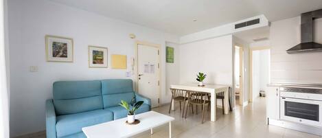 Een moderne woonkamer met een blauwe bank, een witte salontafel en een eethoek met een kleine tafel en stoelen.