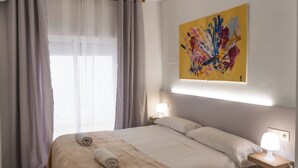 Una habitación de hotel con una cama, dos lámparas de noche, un ventanal con cortinas y un cuadro abstracto de colores en la pared.