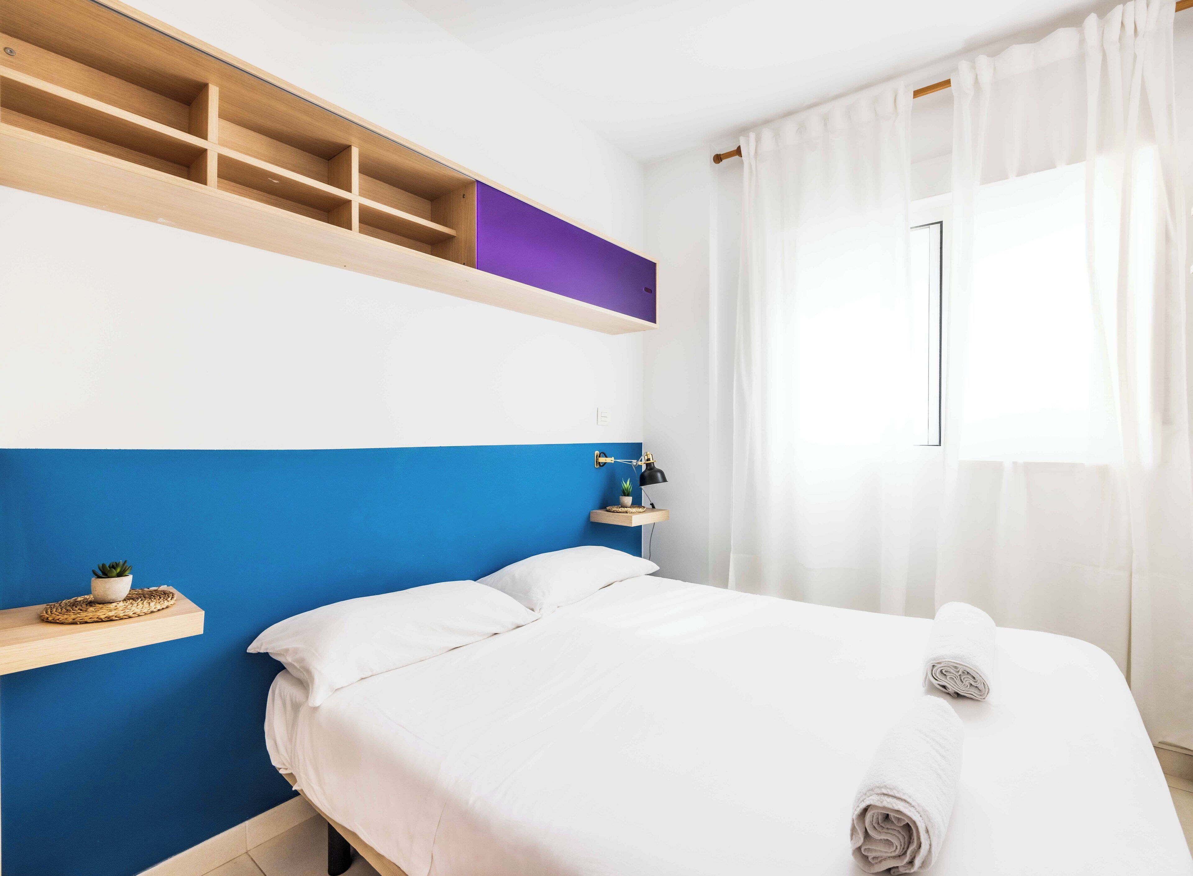 A neatly made bed with white linens, a bedside table with a lamp, and a window with sheer curtains.