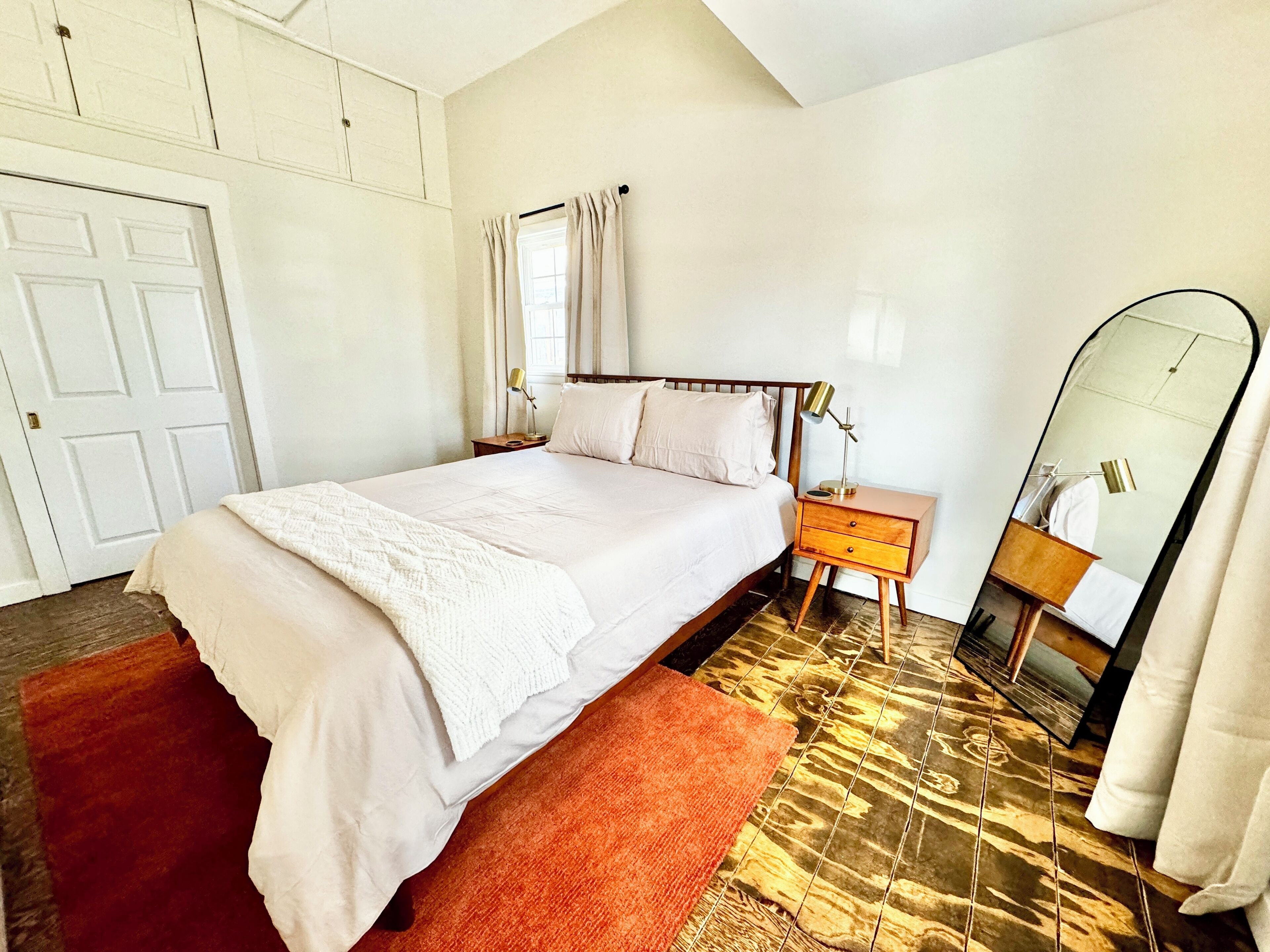 A bedroom with a bed, bedside table, lamp, mirror, and a door.