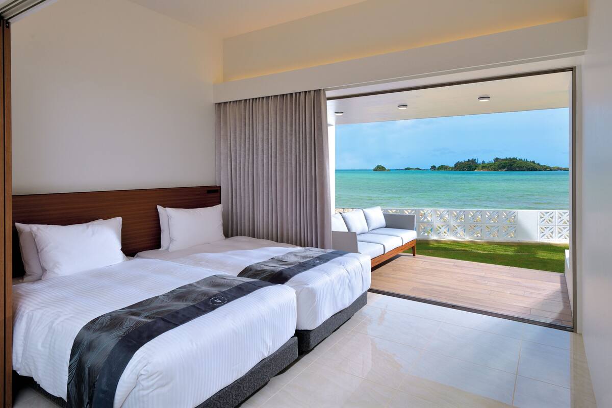 Habitación de hotel con dos camas, un balcón con vista al mar y un cabecero de madera.
