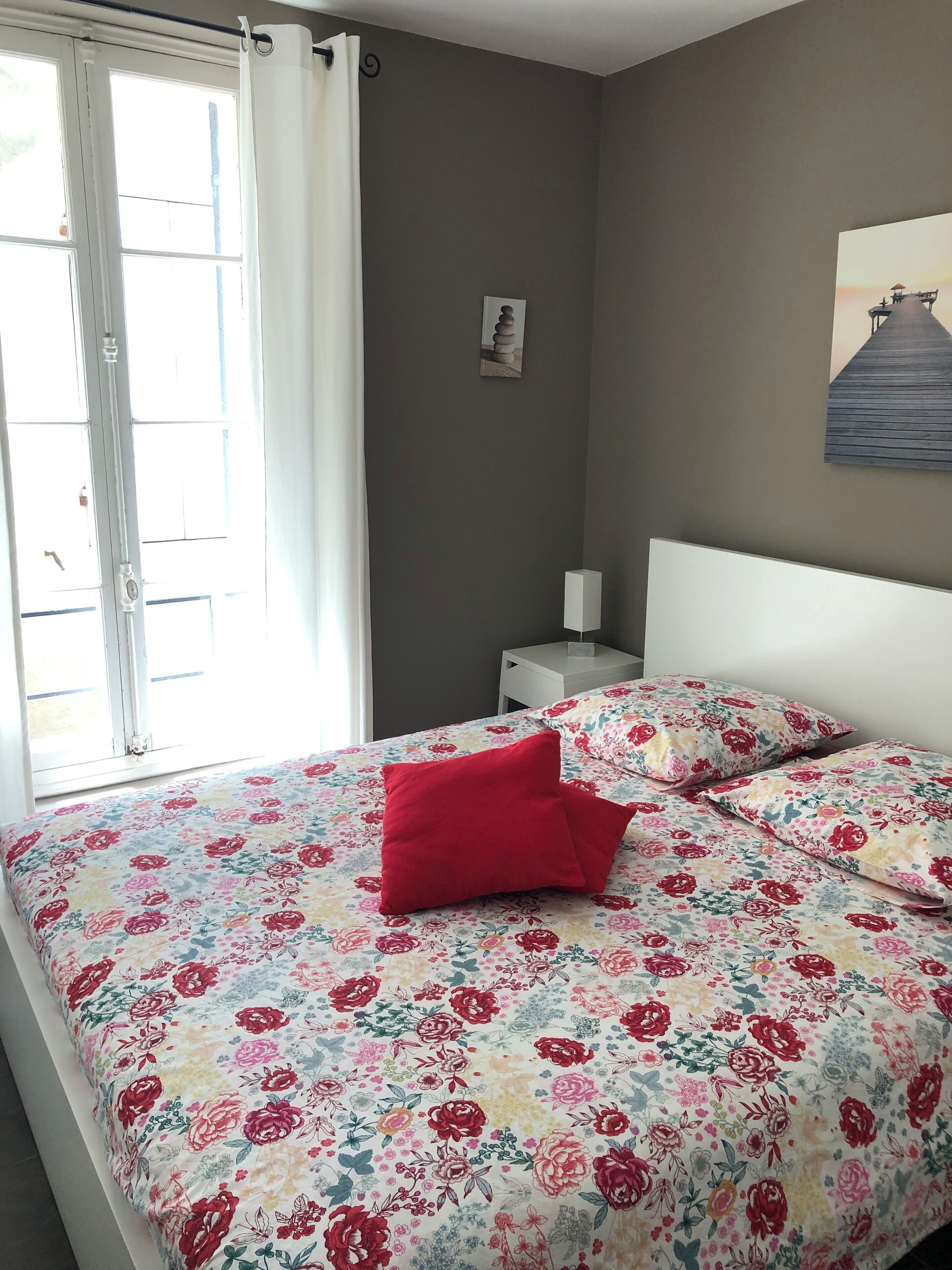 Un dormitorio con cama, mesita de noche, lámpara, ventana con cortinas e imagen en la pared.