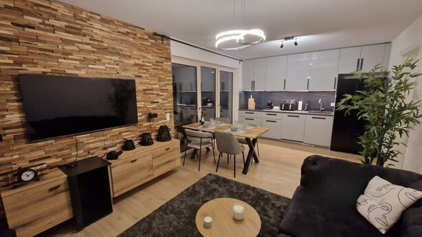 A modern living room with a flat-screen TV, wooden wall, dining area, kitchen, and a plant. - Mr. Grey by the lake (Zeuthen)