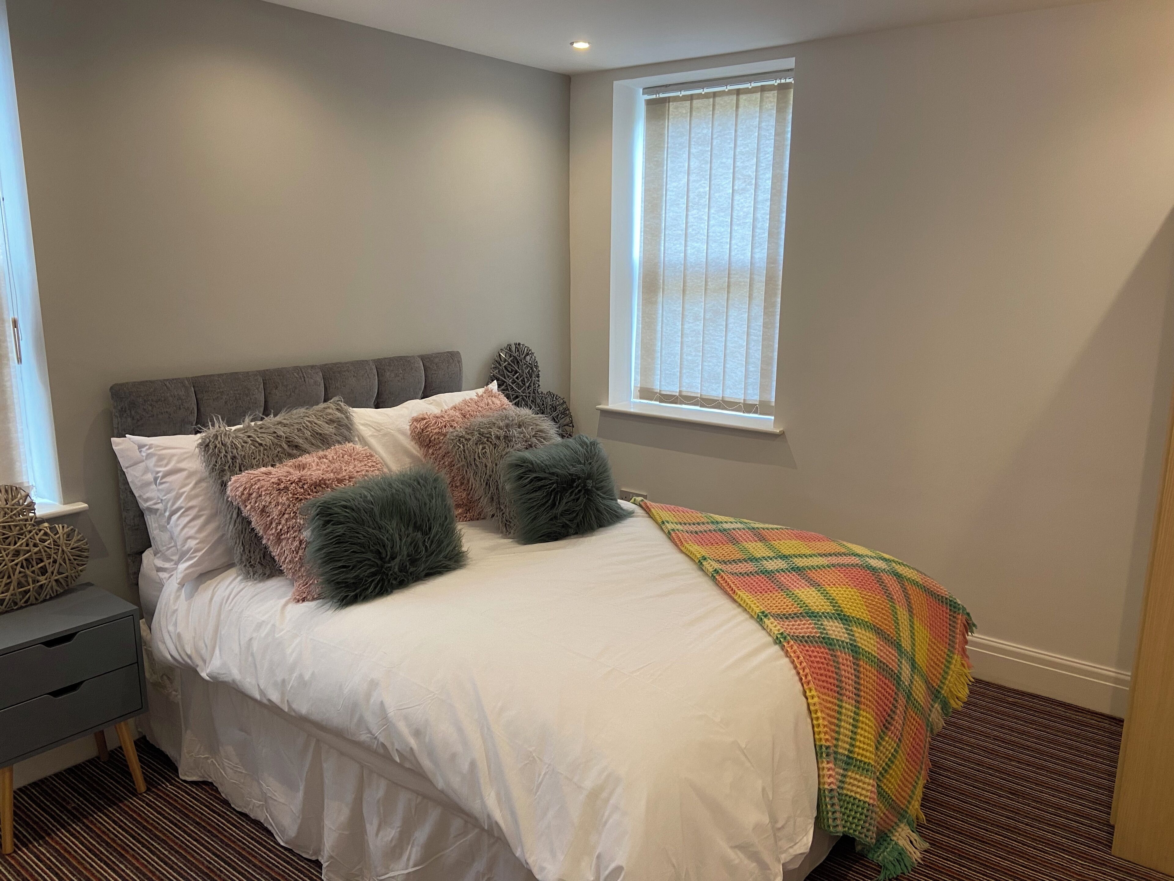 Een slaapkamer met een bed, een grijs hoofdbord, wit beddengoed, zachte kussens, een kleurrijke deken, een nachtkastje en een raam met jaloezieën.