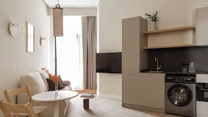 Et moderne køkken med integreret vaskemaskine og tørretumbler, et spiseområde med et rundt bord og stole, samt en opholdsstue med sofa og stort vindue.