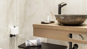 Un baño moderno con pared de mármol, un espejo redondo, tocador de madera, un lavabo grande tipo cuenco y un inodoro.