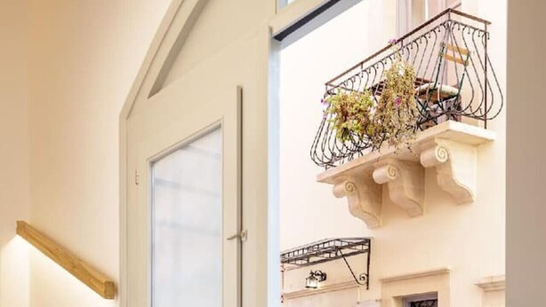 Una escalera conduce a una puerta con un panel de vidrio esmerilado, un balcón con barandilla de hierro forjado y flores, y una calle empedrada al exterior.