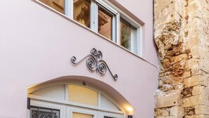 A two-story building with a pink facade, a stone wall on the right, and a double door with decorative metalwork. - Arch.4 Luxury Living (Chania)