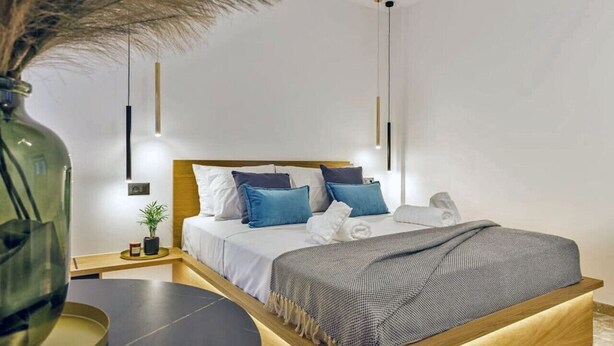 A modern bedroom with a wooden headboard, a bedside table, a nightstand, a large vase, and a hanging light fixture. - Arch.4 Luxury Living (Chania)