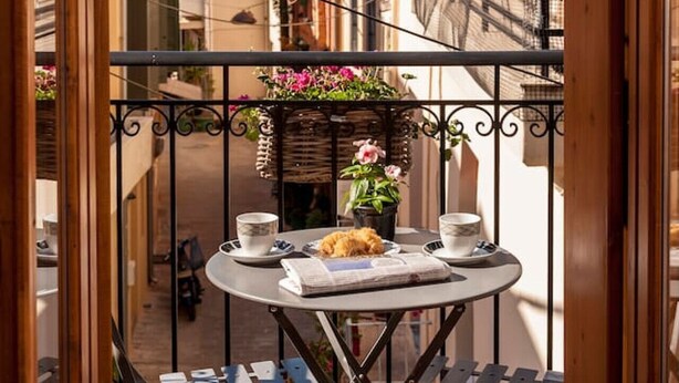 Ein schmaler Balkon im Freien mit einem Tisch für zwei Personen, auf dem sich eine Zeitung, eine Tasse und ein Teller mit Essen befinden.