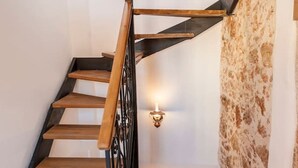 Eine Wendeltreppe mit hölzernen Stufen und schwarzen Metallgeländern, die zu einer Zwischenebene mit einer Steinmauer und einer wandmontierten Leuchte führt.
