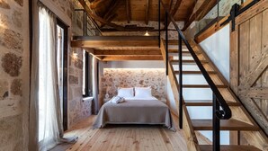 A bedroom with a wooden floor, a bed with a striped bedspread, a stone wall, and a wooden staircase. - Delta-Rho | Kalami | Crete (Kalami)