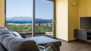 A room with a sofa, a TV, and a sliding glass door leading to a balcony with a view of the sea and buildings.