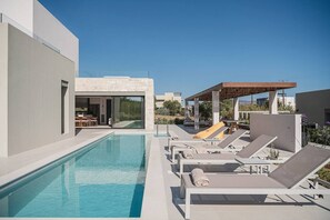 A modern outdoor pool area with lounge chairs, a pergola, and a building with a glass door.