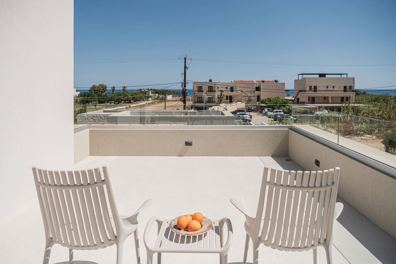 A balcony with two white chairs and a table with a bowl of fruit. The view includes a residential area with buildings and a clear sky.