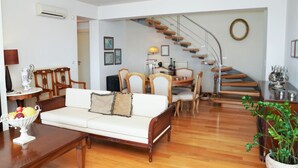 A living room with a white sofa, wooden coffee table, and a fruit bowl. There is a staircase leading to an upper floor, a dining area with wooden chairs, and a potted plant. - Casa Minervos | Unique Views (Souda)