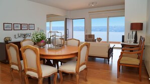 A dining area with a round wooden table and six chairs, a view of the sea through large windows, and a cozy seating area with a sofa and a small table. - Casa Minervos | Unique Views (Souda)