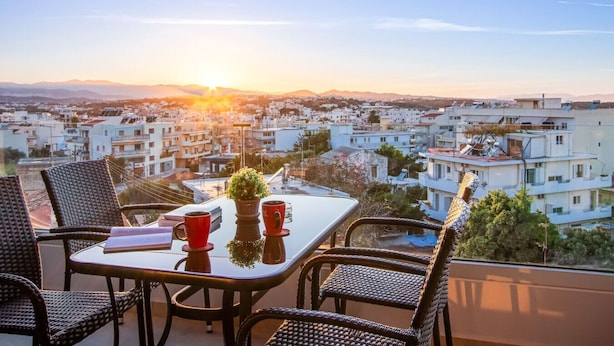 Une terrasse avec des chaises en osier, une table et une vue sur le paysage urbain au coucher du soleil.