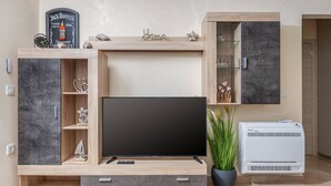 Un salon moderne doté d’un téléviseur à écran plat, de meubles en bois, d’une petite étagère ornée d’objets décoratifs et d’une plante en pot.