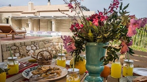 An outdoor dining area with a stone wall, a table set with plates of food, a large vase of flowers, and a view of a building with a tiled roof. - Villa Geraniotis | Chania (Gerani)