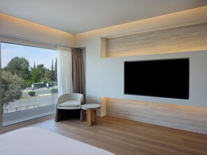 A modern hotel room with a large window, a flat-screen TV mounted on the wall, a comfortable chair, and a small round table. - Nur Riviera Aparthotel (Palaio Faliro)