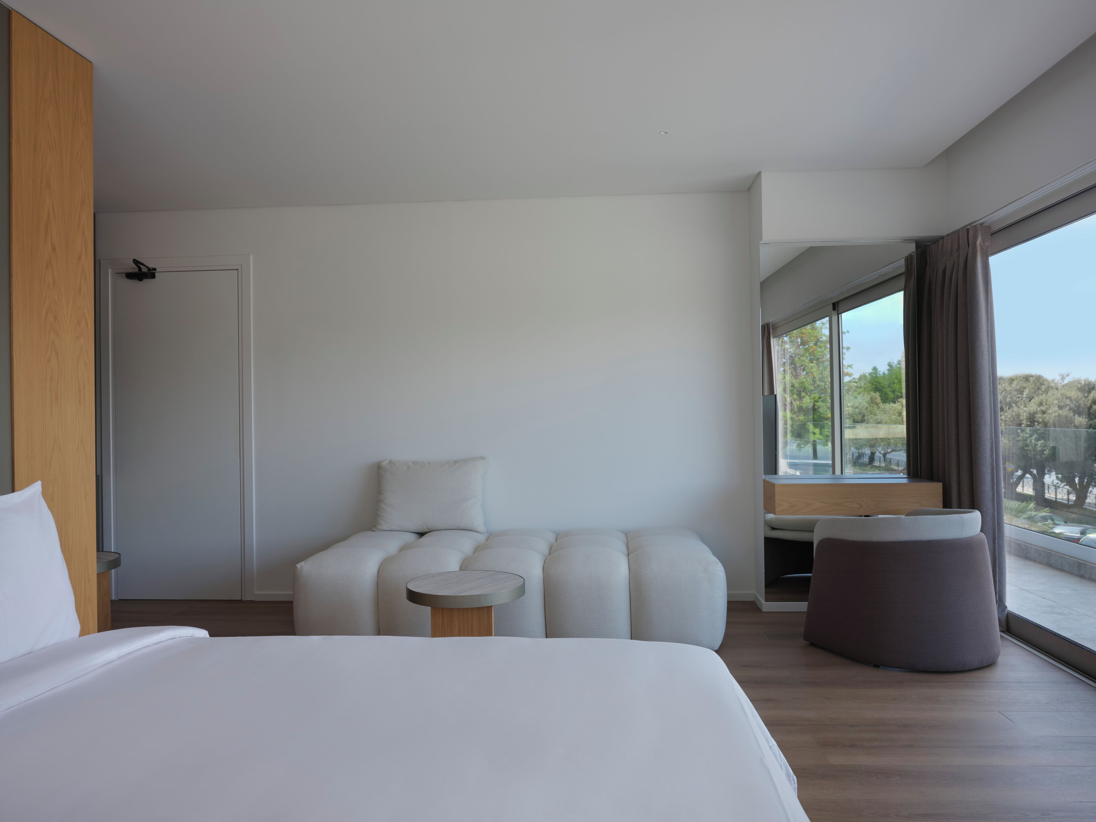 Una camera d'albergo moderna con un letto grande, una poltrona, un tavolino e vista sull'esterno attraverso un'ampia finestra.