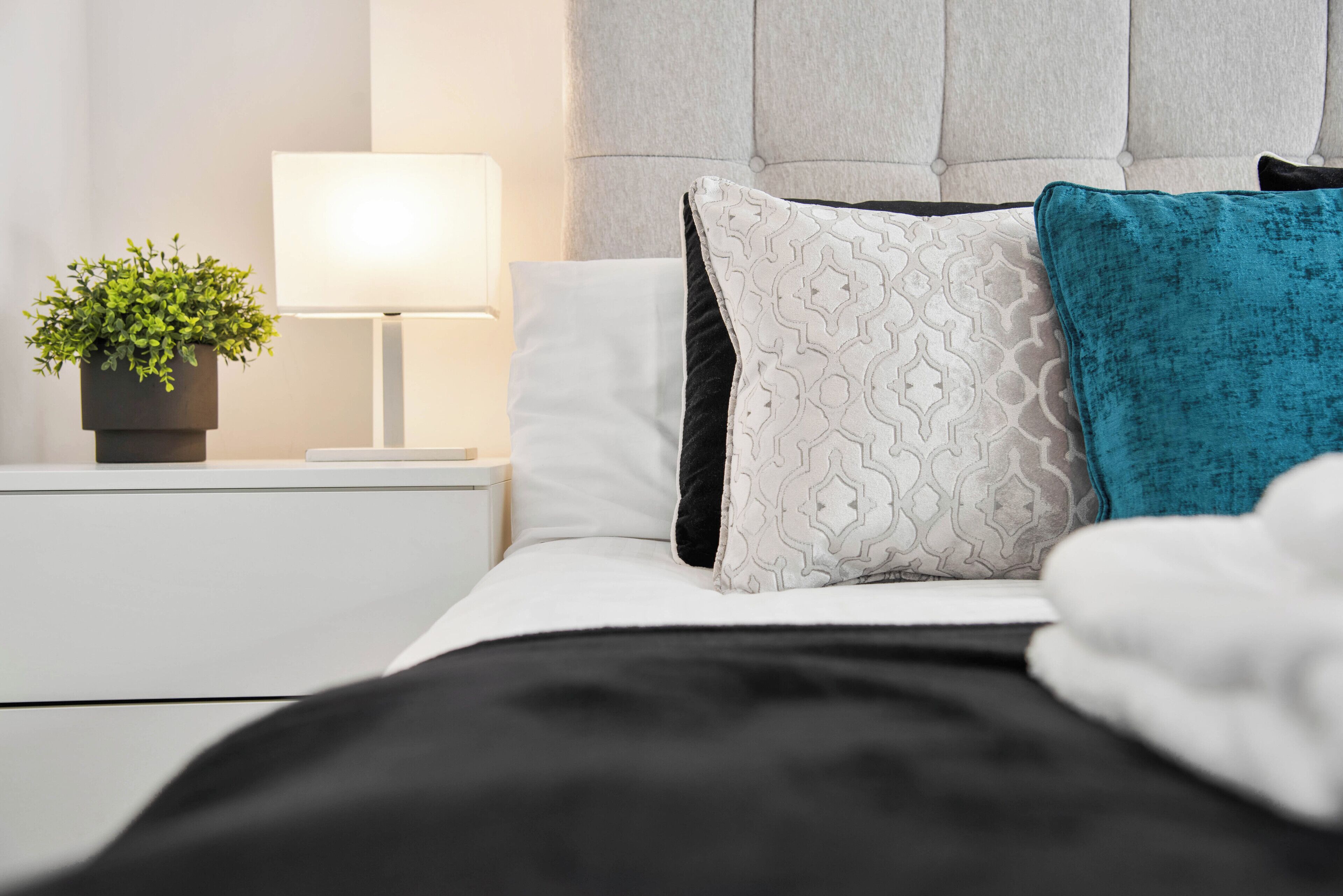 Una cama bien hecha con almohadillas estampadas, una planta en la mesita de noche y una lámpara de noche.