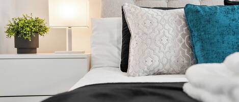 Una cama bien hecha con almohadillas estampadas, una planta en la mesita de noche y una lámpara de noche.