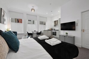 Ett modernt hotellrum med en stor säng, ett skrivbord, ett litet kök och en tv som är monterad på väggen.