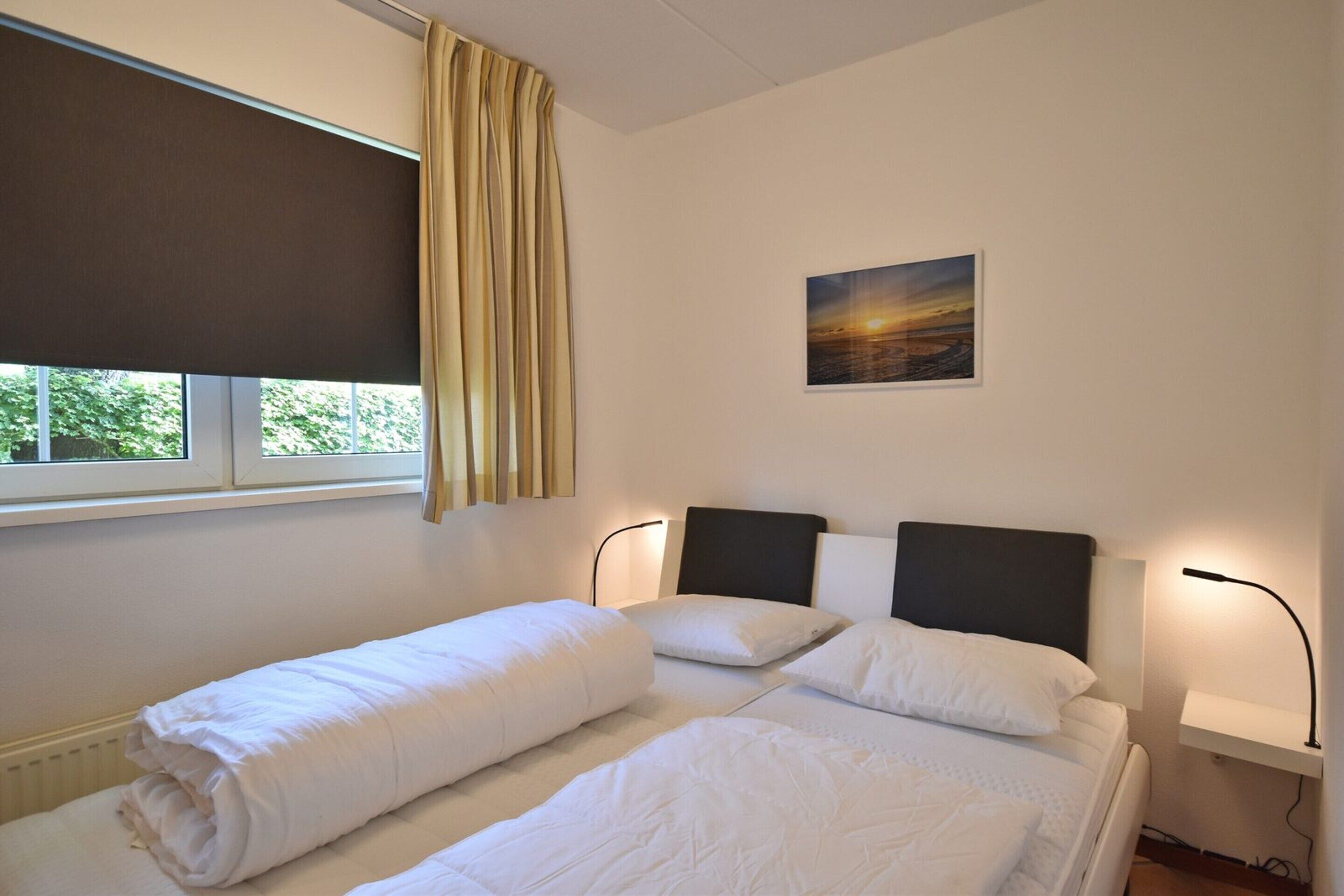A hotel room with two beds, a window with a view, a painting on the wall, and a bedside table with a lamp.