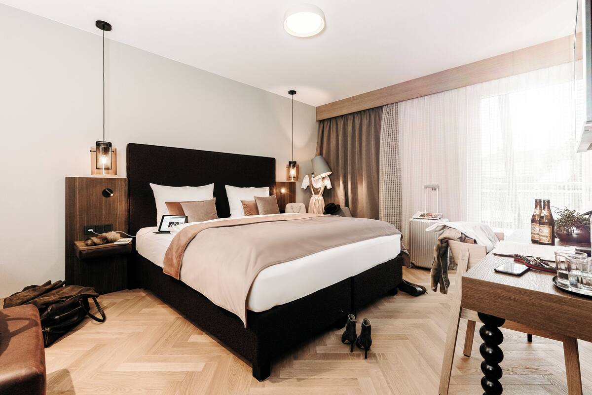 Una habitación de hotel moderna con una cama grande, mesitas de noche, un escritorio y vistas a la ciudad desde la ventana.