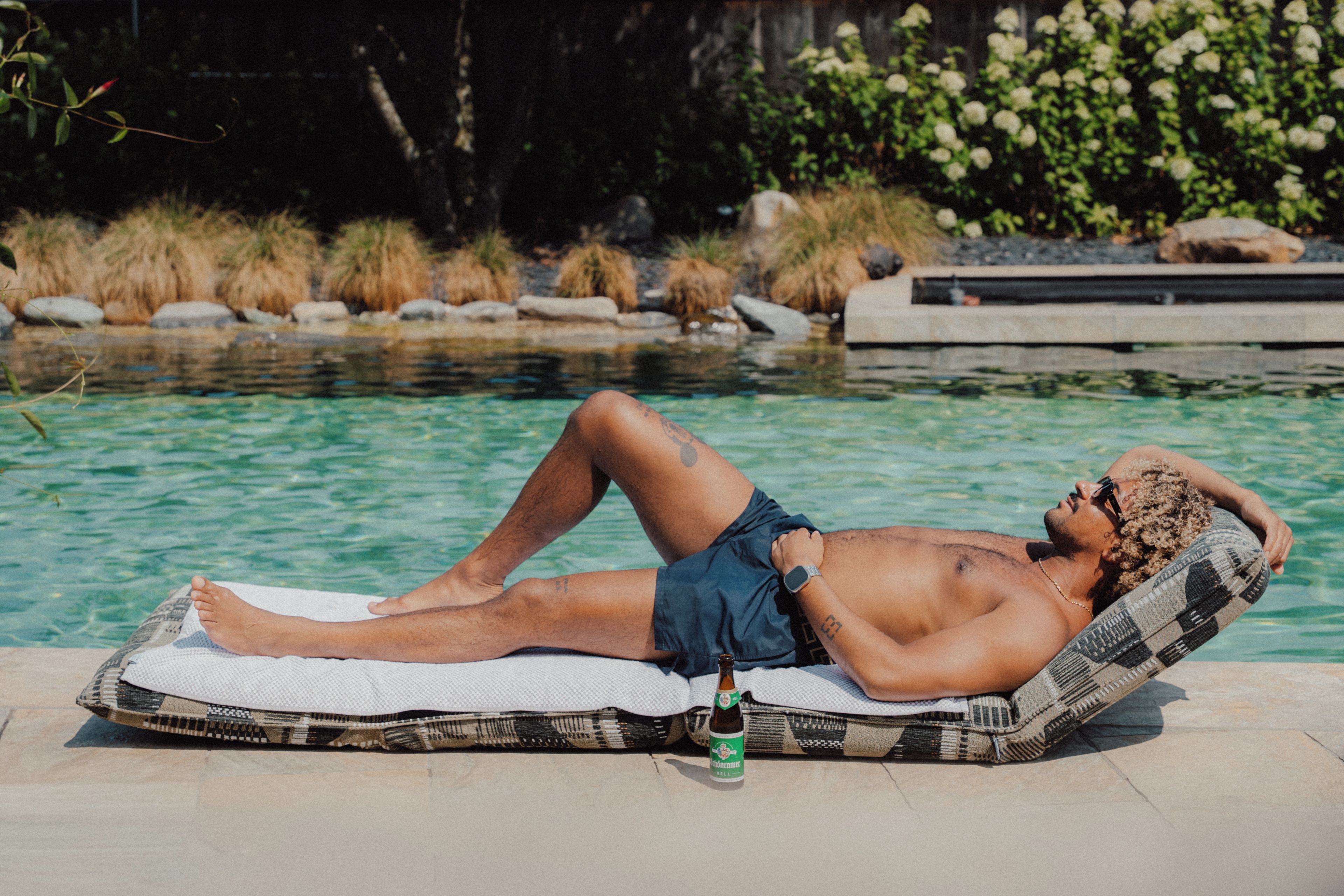A person lying on a lounge chair by a pool with a bottle of beer.