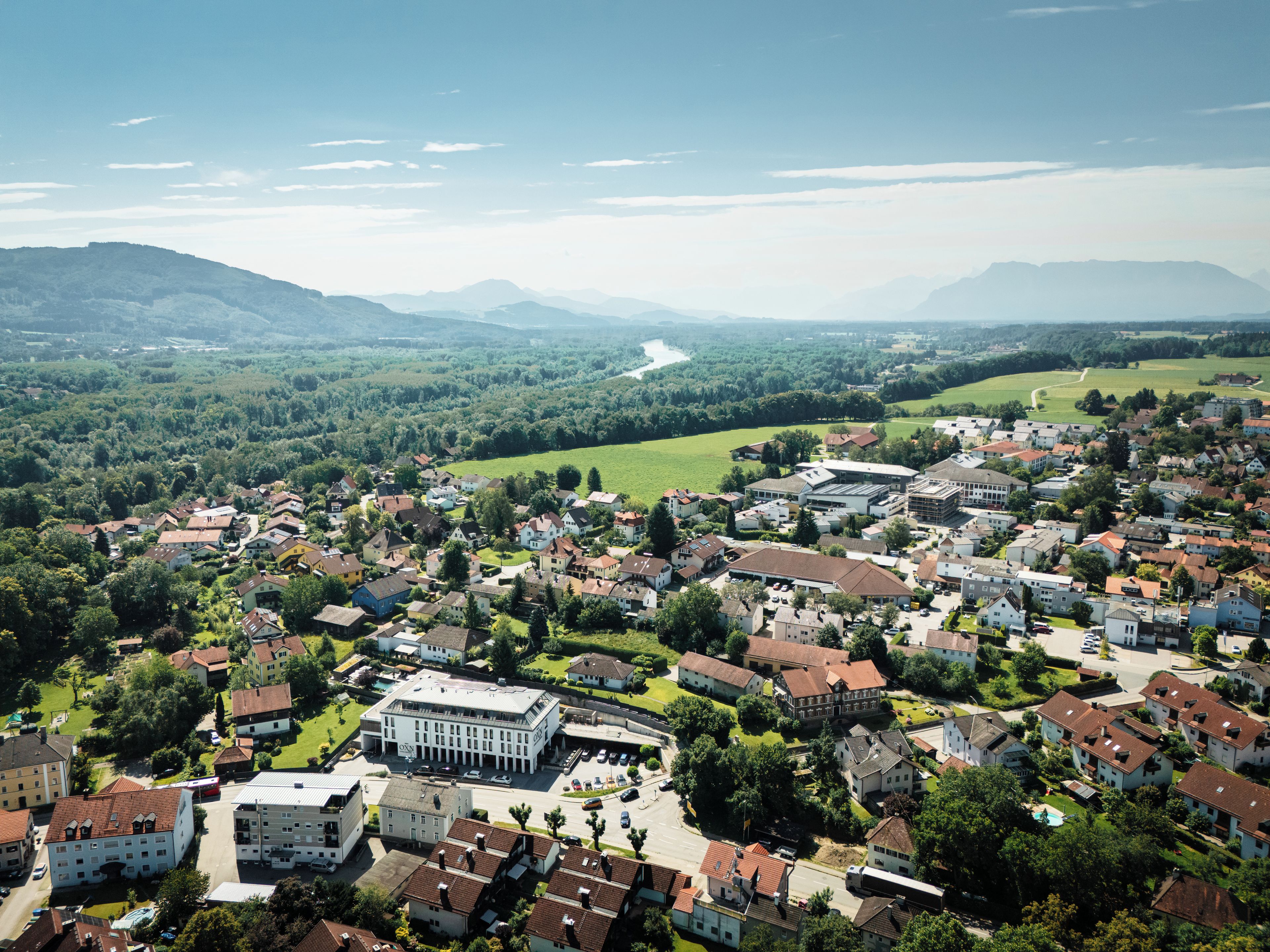 An aerial view of a town with a mix of residential and commercial buildings, surrounded by greenery and mountains.