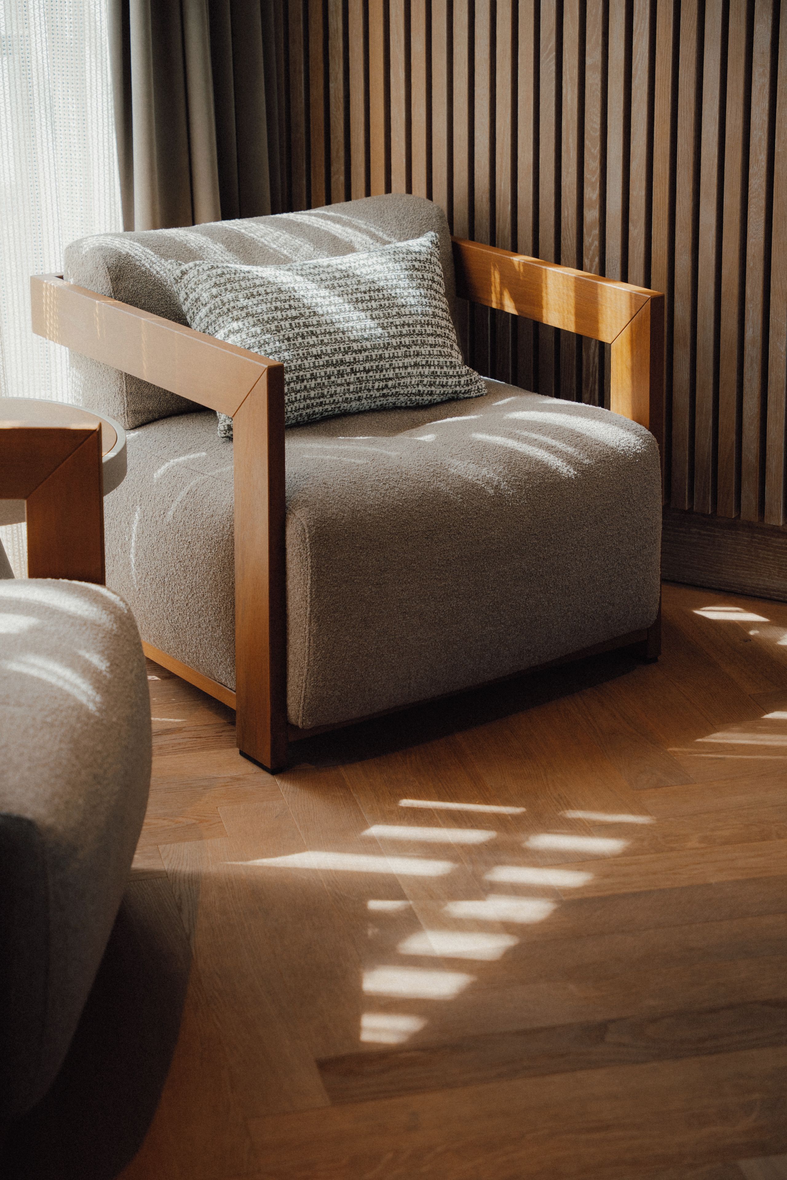 Ein moderner Stuhl mit Holzrahmen und grau gepolsterter Sitzfläche, platziert in einem Raum mit Holzboden und einer Wandpaneele aus Holz.