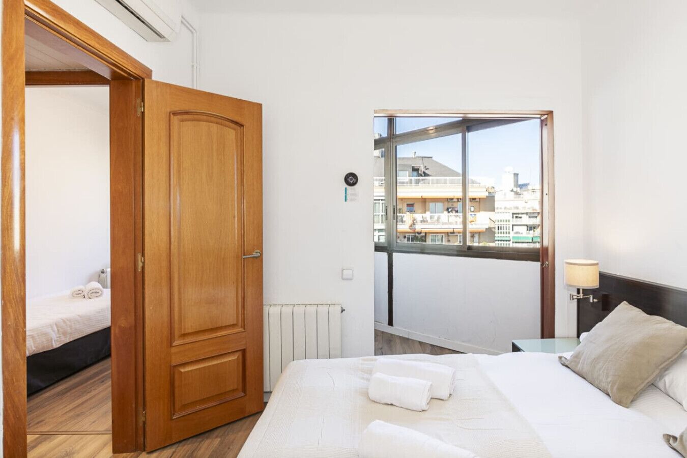 Ein Hotelzimmer mit Holztür, einem Bett mit weißer Bettwäsche, Blick auf Gebäude durch ein großes Fenster und einer Nachttischlampe.
