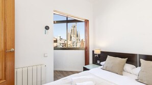 A hotel room with a large bed, a nightstand, a window with a view of a cityscape, and a wooden door. - Vistas SAGRADA FAMILIA piso grande y luminoso (Barcelona)