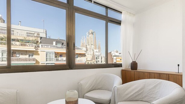 A modern room with two white armchairs, a small round table, and a view of a cityscape through large windows. - Vistas SAGRADA FAMILIA piso grande y luminoso (Barcelona)