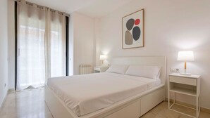 A modern hotel room with a large bed, a bedside table with a lamp, a radiator, and a window with curtains. - Apartamento para 6 amplio y luminoso en Bonanova (Barcelona)