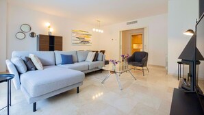 A modern living room with a grey sectional sofa, a glass coffee table, and a dark armchair. There is a painting on the wall and a small table with flowers. - Apartamento para 6 amplio y luminoso en Bonanova (Barcelona)
