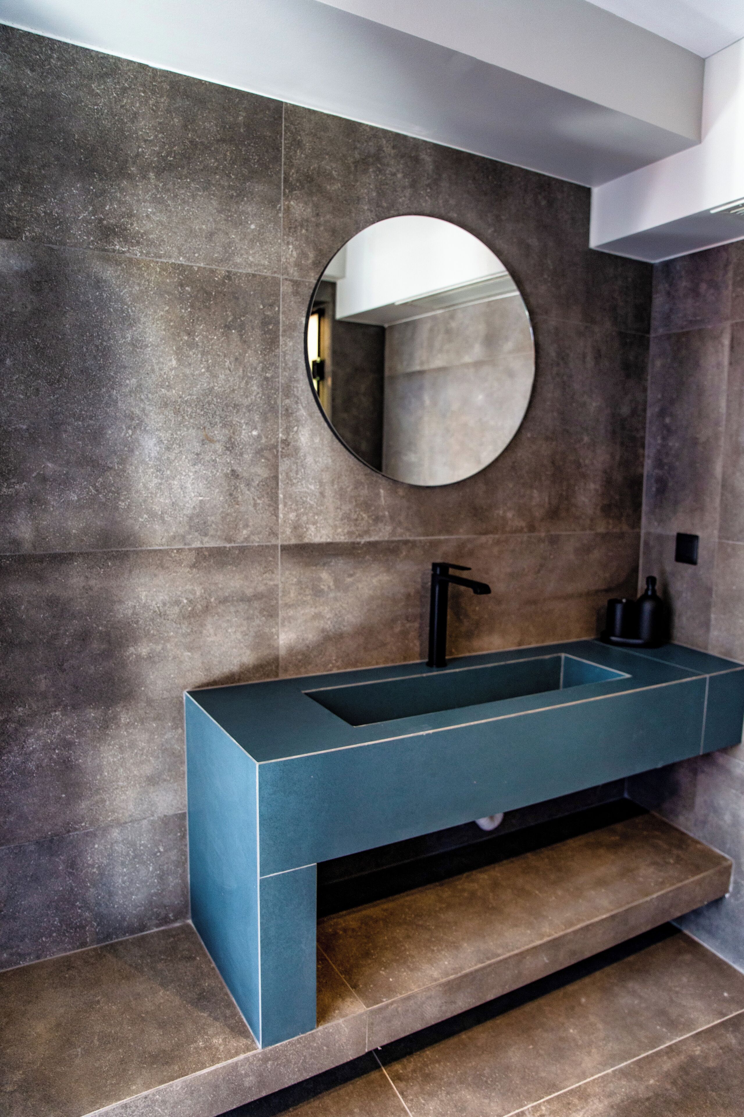 Un bagno moderno con un lavabo rettangolare, uno specchio rotondo e piastrelle in pietra scura.