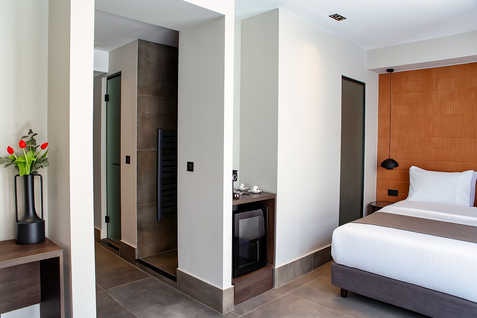 A modern hotel room with a bed, bedside table, and a small desk with a chair.