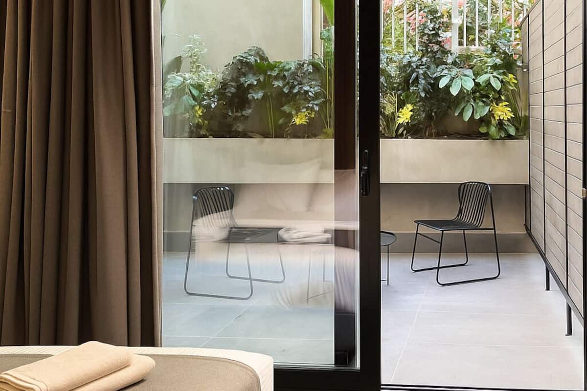 A modern bedroom with a bed, a sliding glass door leading to a balcony with plants and a chair.