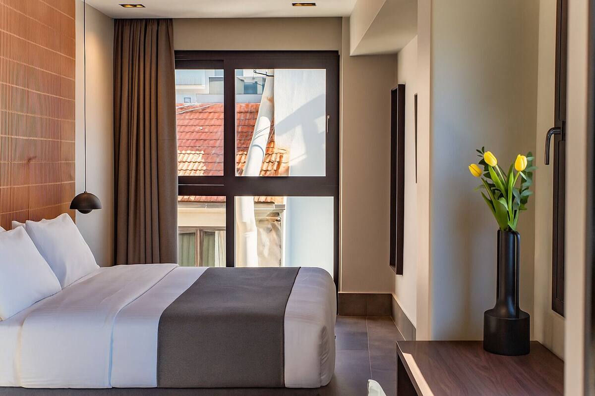 A modern hotel room with a large bed, a view of the cityscape, and a vase of yellow flowers.