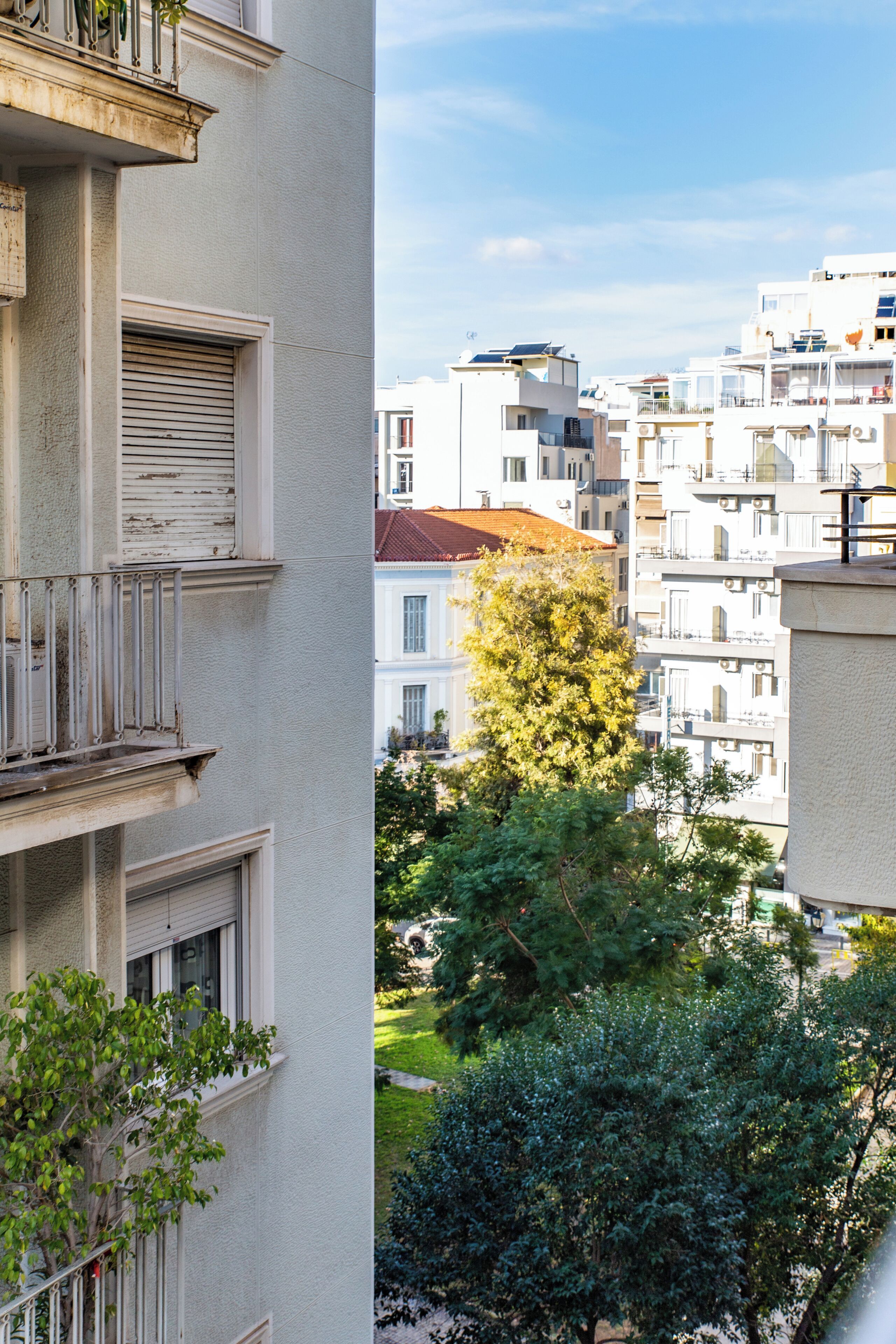 A multi-story building with balconies, a tree in the foreground, and other buildings in the background.