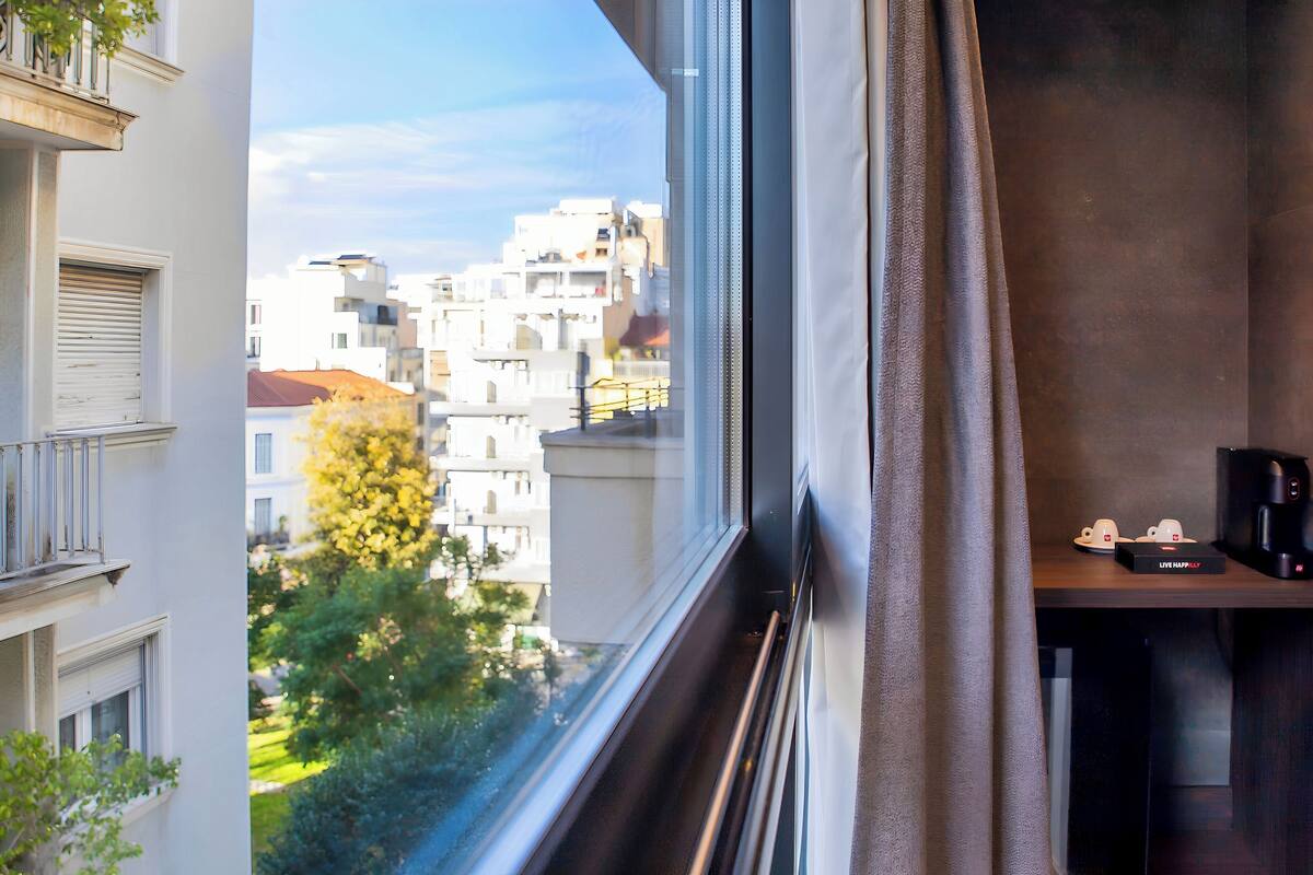 A room with a view of neighboring buildings, a window with blinds, a small table with a coffee maker, and a curtain.