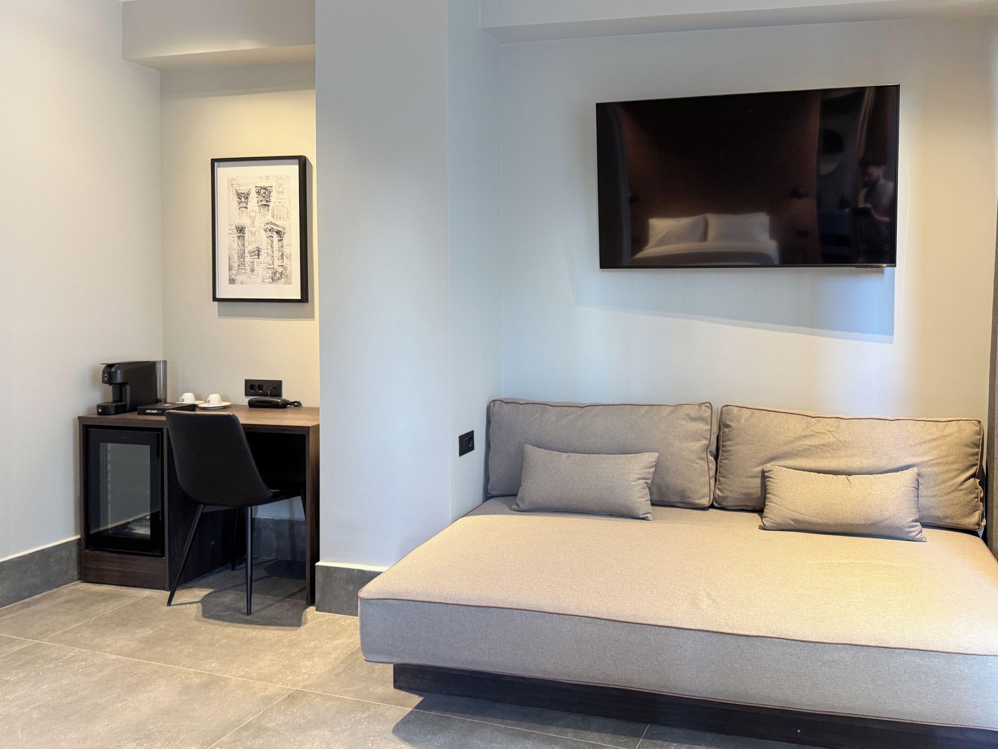 A modern room with a bed, a desk, and a chair. There is a framed picture on the wall and a flat-screen TV mounted above the bed.