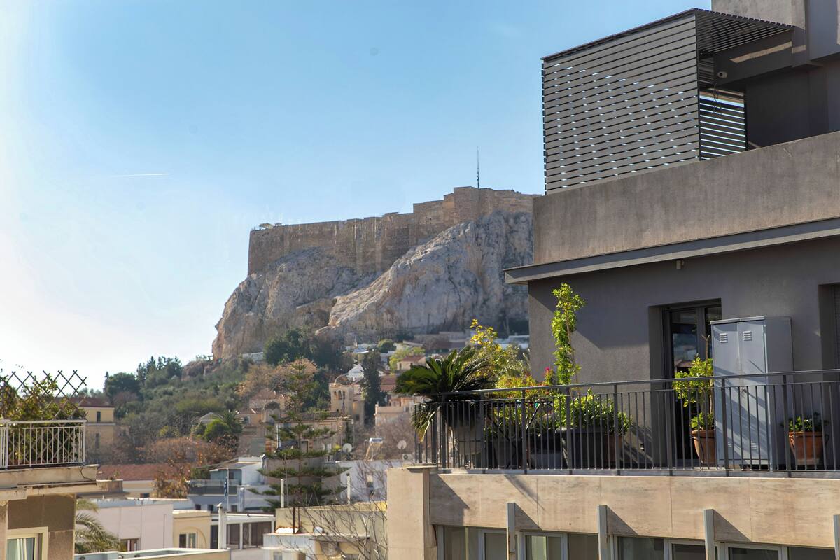 A modern building with a balcony, potted plants, and a view of a rocky hill with a castle-like structure in the background.