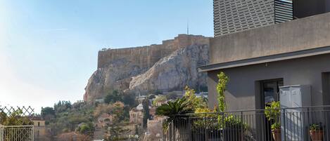 A modern building with a balcony, potted plants, and a view of a rocky hill with a castle-like structure in the background.